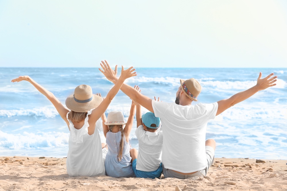 Family with two adults and three kids sits on the beach facing the ocean with arms raised in joy.