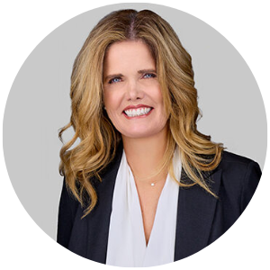 Professional headshot of a smiling woman with blonde hair, wearing a navy blazer and white blouse, against a light gray background.
