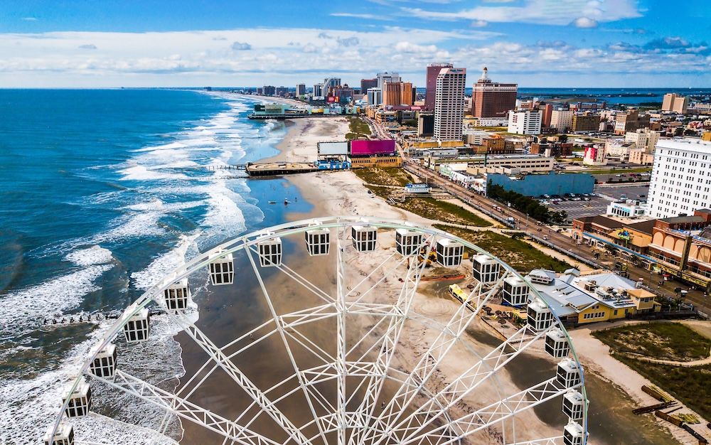 Aerial view of a coastal city with a large white Ferris wheel along the sandy shoreline and blue ocean on the left.”,