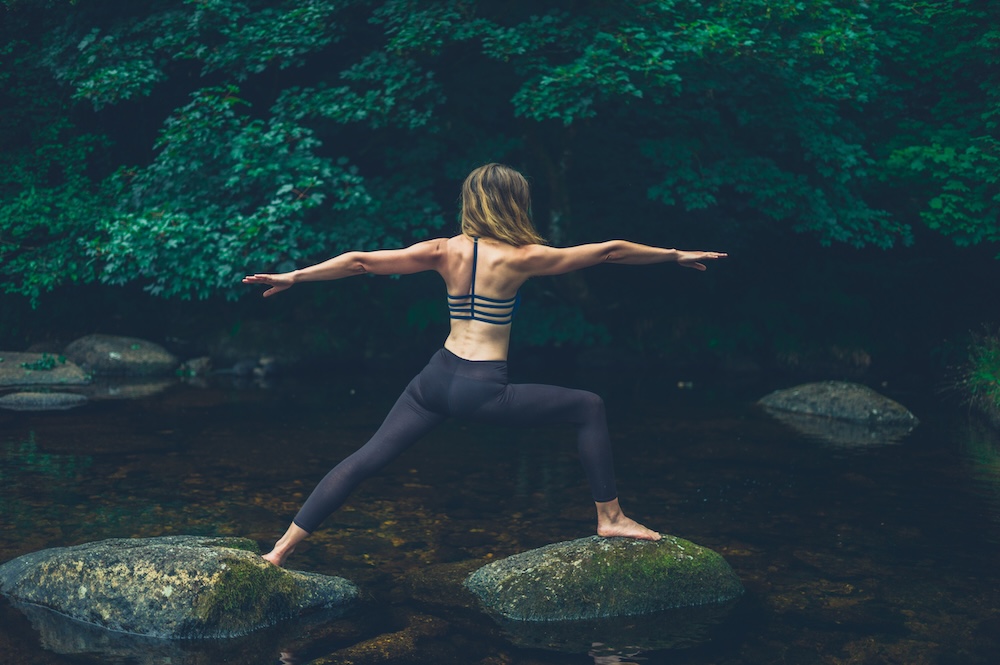 Woman practicing yoga on a mossy rock in a shallow stream, arms extended in a balance pose with a forest backdrop beyond.