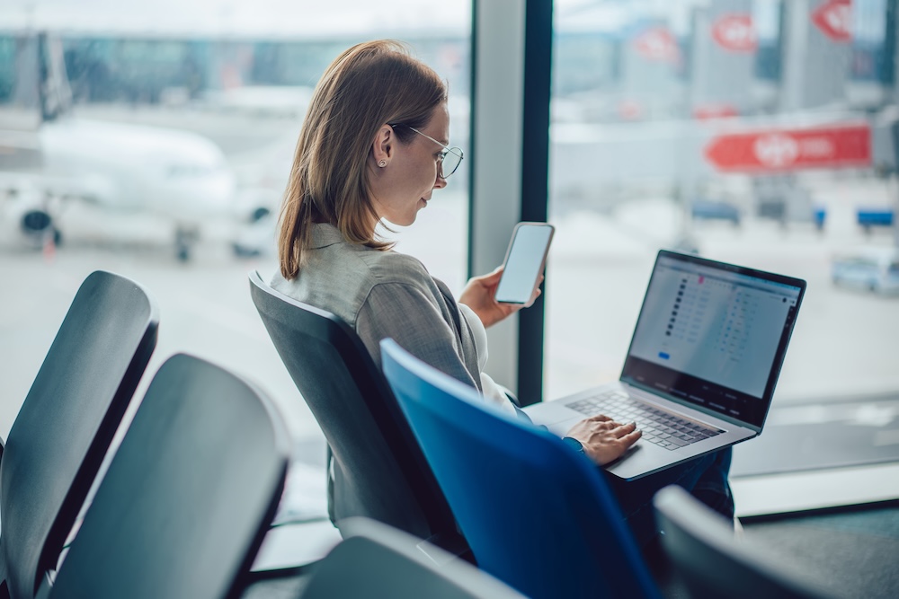 Side view of female passenger on smartphone and using touchpad on laptop to surf internet while sitting at airport against blurred airfield